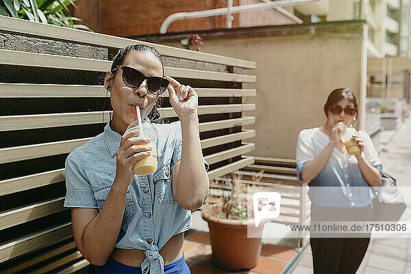 Female friends wearing sunglasses drinking juice while standing in city
