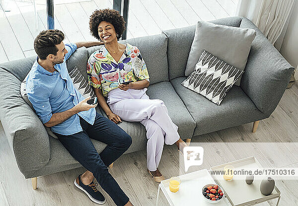 Happy multi-ethnic couple holding smart phones while sitting on sofa in penthouse