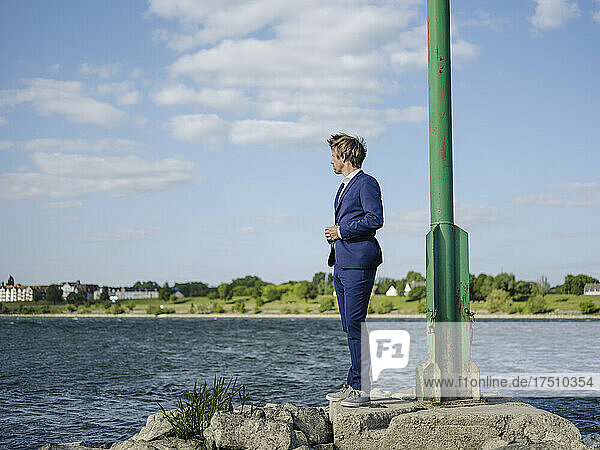 Businessman wearing suit standing on rock at riverbank against sky