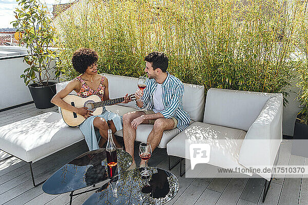 Glückliche Frau spielt Gitarre für Mann und trinkt Rotwein auf dem Sofa auf der Penthouse-Terrasse