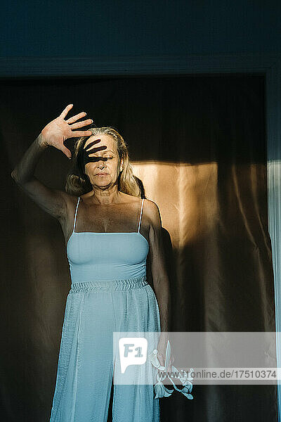 Senior woman shielding face from sunlight with hand while standing against curtain