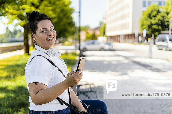 Portrait of a smiling curvy young woman with mobile phone sitting on a bench in the city