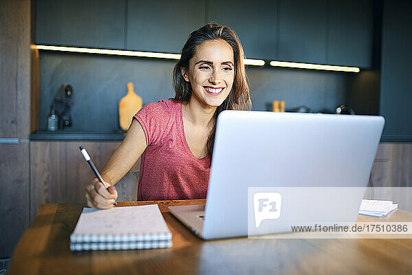 Smiling female entrepreneur looking at laptop while writing in note pad on desk