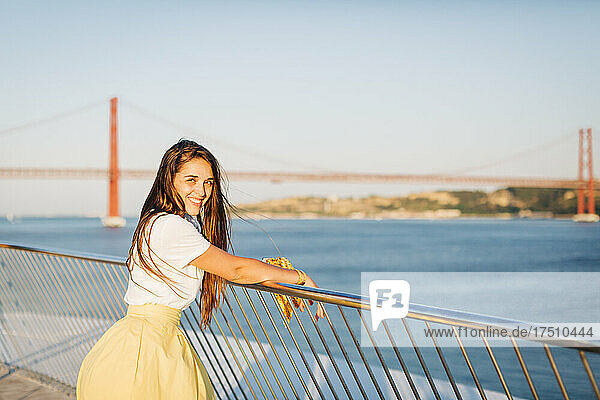 Smiling beautiful woman with long hair standing by railing over river in city at sunset