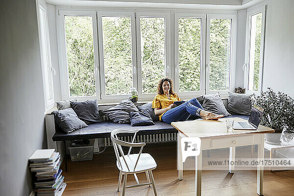 Woman with digital tablet relaxing on sofa against window at home