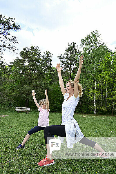 Mother with daughter practicing warrior 1 pose on grassy land in forest
