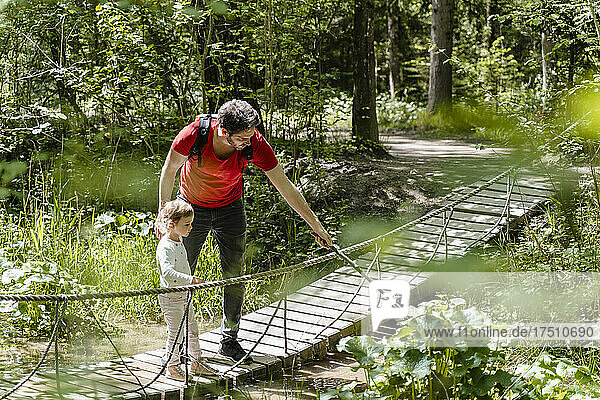 Father and daughter looking at plants while standing on footbridge in forest