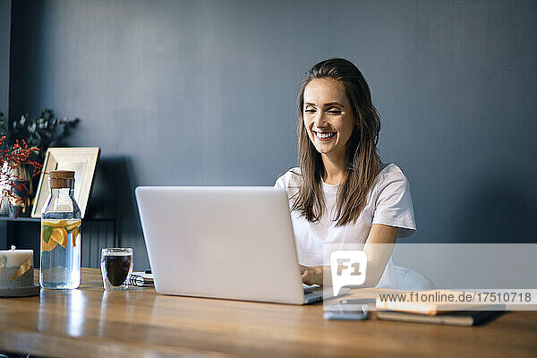 Smiling young woman using laptop on desk against wall in home office