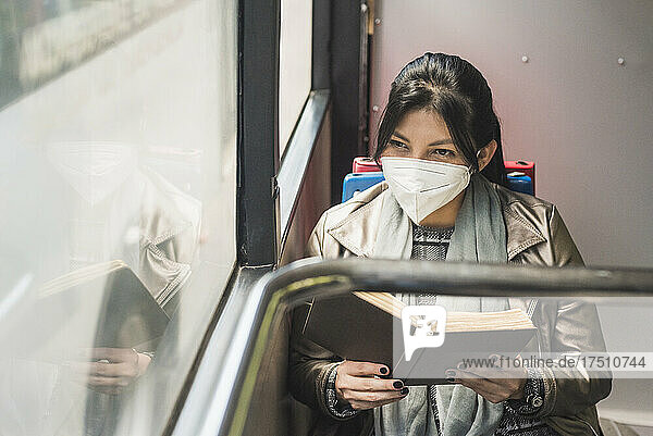 Woman wearing protective mask  looking through window while reading book in bus