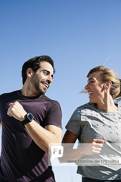 Smiling couple looking at each other while running against clear blue sky