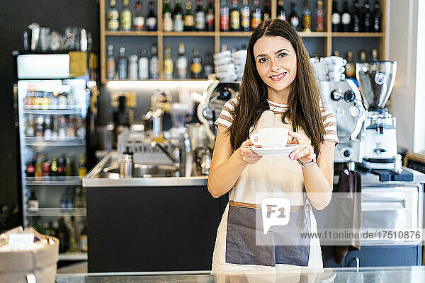 Beautiful young owner holding coffee cup and saucer while standing in cafe