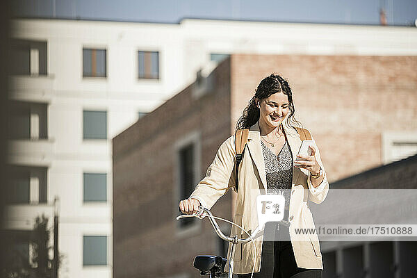 Smiling female commuter using mobile phone while walking with bicycle against buildings in city