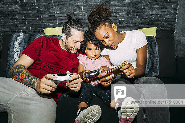 Mother using smart phone while father and daughter playing video game on sofa at home
