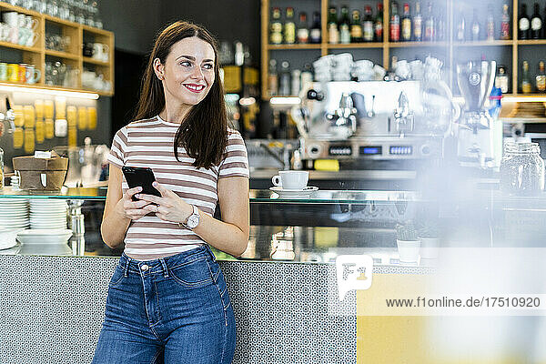 Thoughtful woman holding smart phone while standing at coffee shop