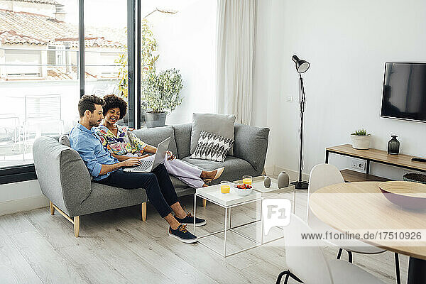 Multiethnisches Paar benutzt Laptop  während es auf dem Sofa in einem modernen Penthouse sitzt
