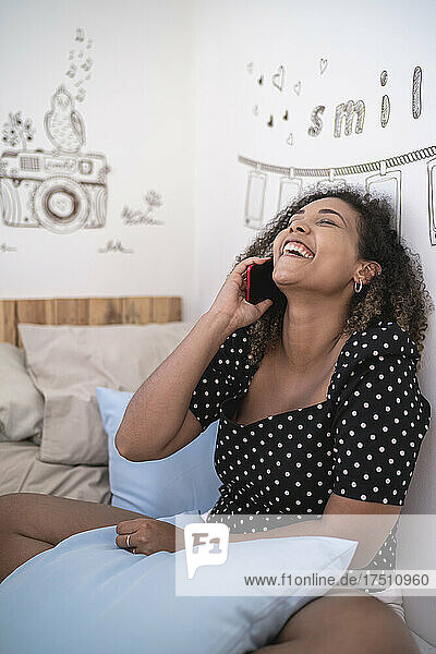 Cheerful woman with curly hair talking over smart phone while sitting against wall at home