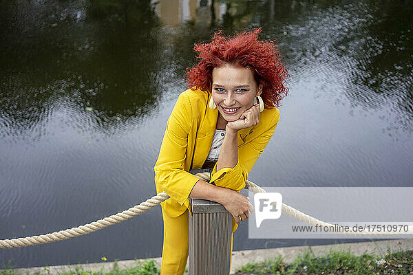 Junge Frau mit lockigem Haar und gelbem Anzug steht am Fluss und stützt sich auf eine Stange