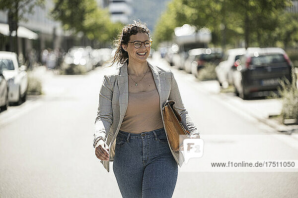 Lächelnde junge Frau mit Tasche  die an einem sonnigen Tag auf der Straße läuft