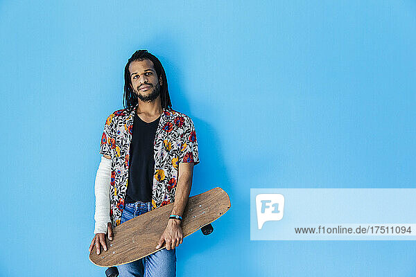 Mitte erwachsener Mann mit gebrochenem Arm hält Skateboard vor blauem Hintergrund
