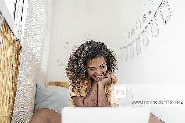 Fröhliche Frau mit lockigem Haar führt Videokonferenzen über Laptop  während sie zu Hause an der Wand sitzt