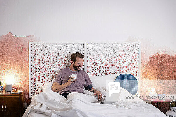Lächelnder erwachsener Mann hält Kaffee mit Laptop in der Hand und entspannt sich zu Hause im Bett