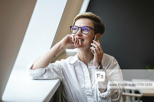 Smiling businesswoman talking on mobile phone while looking through window in office cafeteria