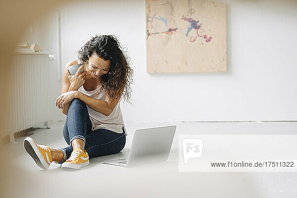 Smiling woman holding coffee mug sitting with laptop on floor at home