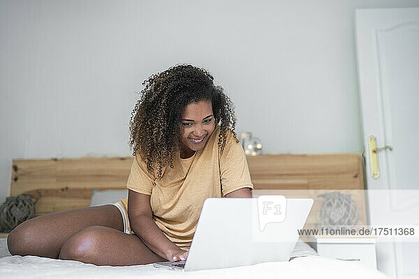 Lächelnde junge Frau mit lockigem Haar benutzt Laptop  während sie zu Hause im Bett sitzt