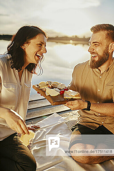 Happy couple having picnic on jetty at a lake at sunset eating cheese