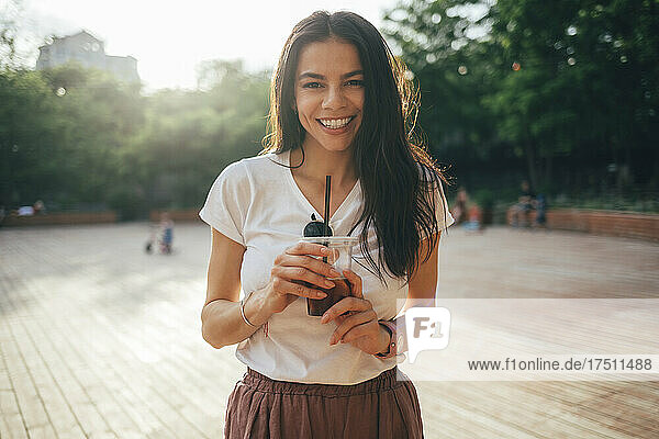 Cheerful beautiful woman with long hair holding soft drink while standing in park