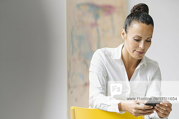 Female entrepreneur using mobile phone while sitting on chair against wall in home office