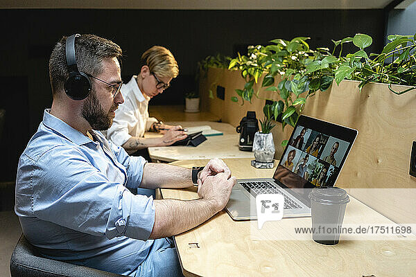 Businessman on video call with colleagues through laptop while sitting by businesswoman at desk in creative office