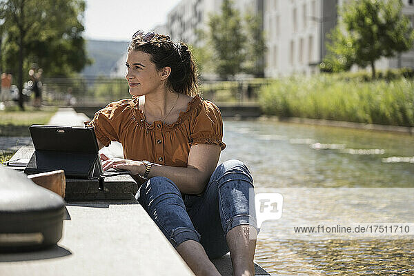 Thoughtful woman with laptop sitting by pond in park during sunny day