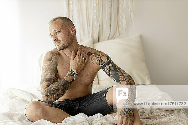 Shirtless hipsterwith tattoo on arms sitting on bed