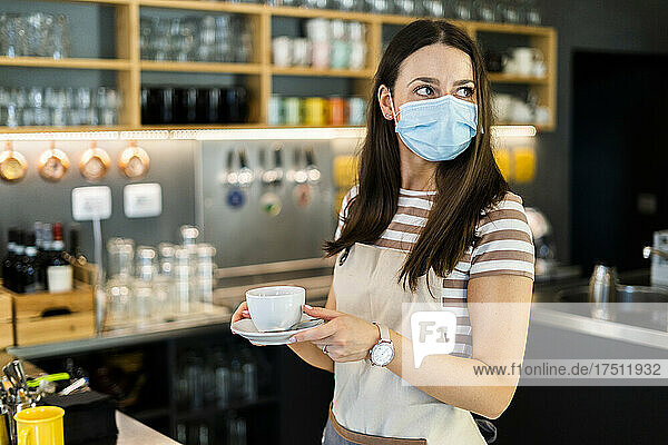 Thoughtful young owner wearing mask while holding coffee cup and saucer in cafe