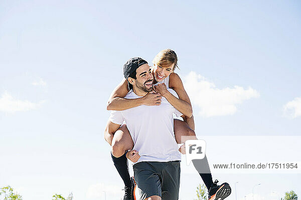 Happy man piggybacking woman while walking against sky during sunny day
