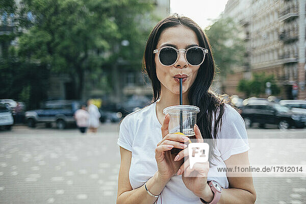 Young woman wearing sunglasses drinking soft drink while standing in city
