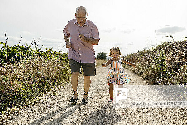 Verspielter älterer Mann rennt mit Enkelin auf unbefestigter Straße inmitten von Pflanzen
