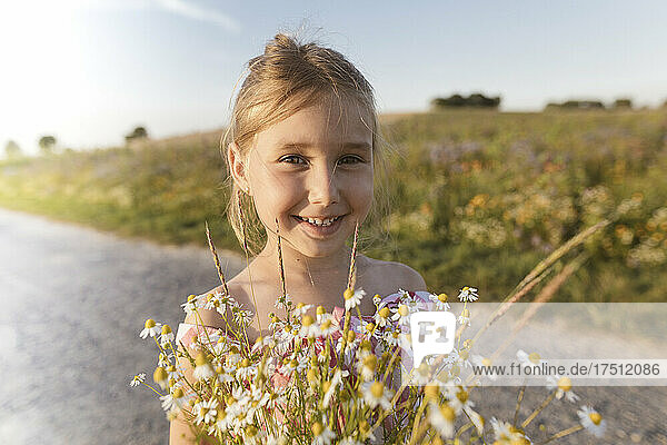 Close-up of smiling girl holding flowers on road against sky at sunset