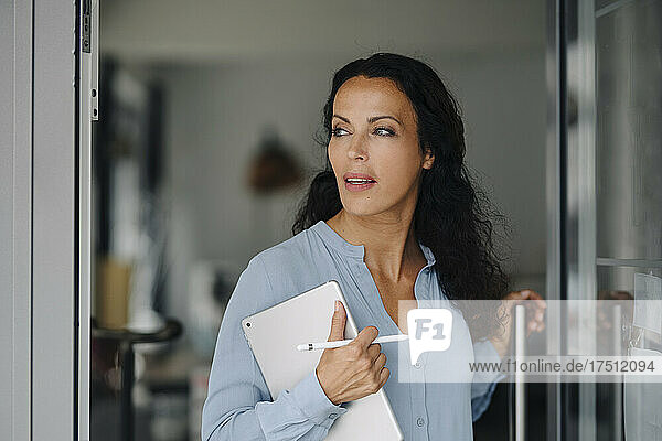 Female owner holding digital tablet looking away while standing at entrance of cafe