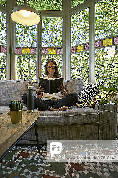 Woman reading book while sitting on sofa in living room