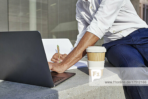 Businessman with laptop and disposable cup writing while sitting in city