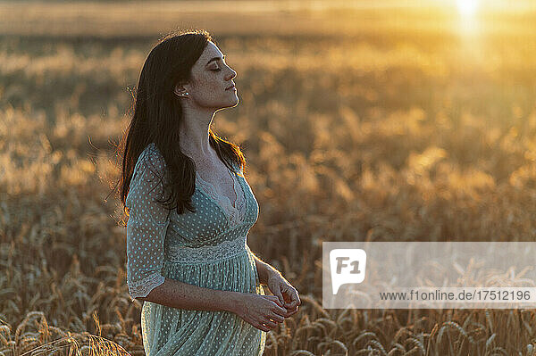 Young woman with eyes closed standing amidst wheat crops in farm at sunset