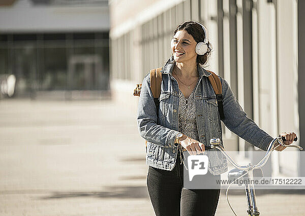 Smiling female commuter listening music while walking with bicycle on street in city