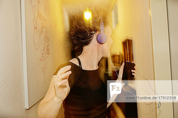 Carefree mid adult woman with headphones dancing at illuminated home