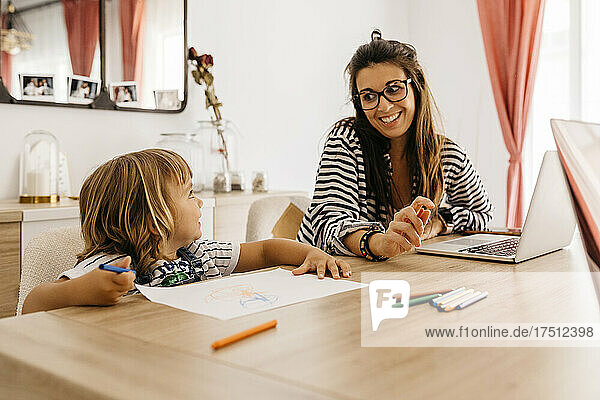Smiling mother with laptop looking at daughter painting on table in dining table