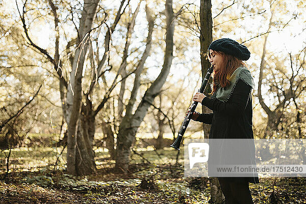 Young female musician practicing playing clarinet while standing in forest during autumn