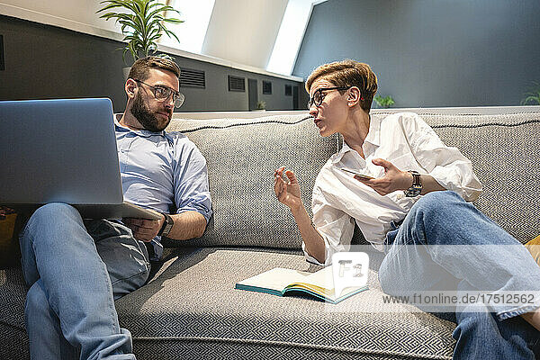 Männliche und weibliche Geschäftskollegen diskutieren  während sie auf einem beleuchteten Sofa in Coworking-Büroräumen sitzen