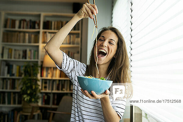 Junge Frau isst Spaghetti  während sie zu Hause am Fenster sitzt