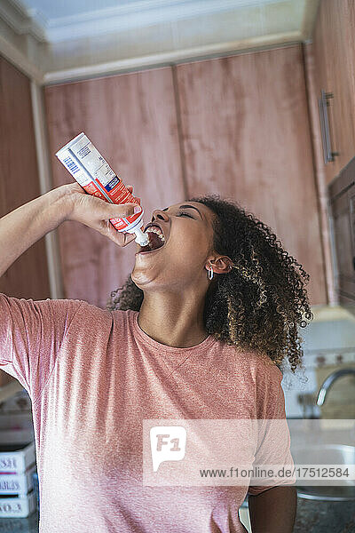 Cheerful young woman with curly hair eating whipped cream while standing in kitchen